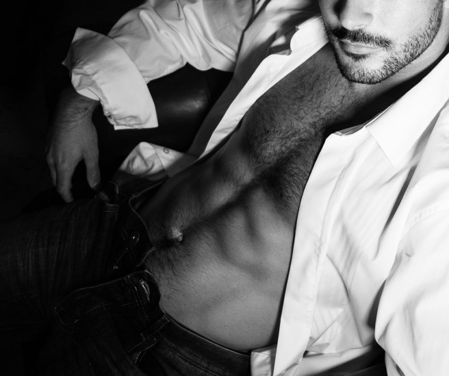 Portrait of attractive young man with beard and open shirt revealing sixpack abs, sitting in leather armchair